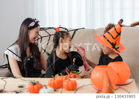 Family celebrates halloween day, two kids in costume are showing a fake bloody hand to their mother, pumpkin buckets and spider decorations are seen on the table. 130433905