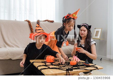 Two children wearing halloween costumes prepare decorations, they put toy pumpkins and a spider web on the table, with an adult nearby watching them. 130433908
