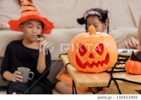 Kids celebrate with pumpkins and spooky decorations for halloween day, one child wears a witch hat while the other holds a fake spider toy indoors. 130433909