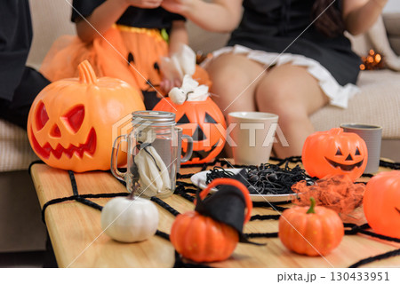 Children in costumes celebrate halloween day with jack o lanterns, spiders and a mason jar on a table decorated with a spiderweb, seated on a couch indoors for the spooky holiday. 130433951