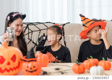 Three kids celebrate halloween day, a girl holds a jar with spiders, another has a black glove and a fake eye, the last one has a witch hat and many pumpkins are on the table. 130433957