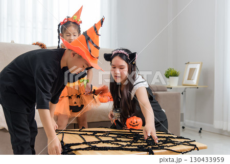 Three children celebrate halloween day, one is wearing a witch hat and dress, another has cat ears while they decorate a wooden table with spider web decor creating a festive atmosphere. 130433959