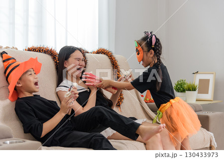 Family celebrates a fun Halloween together, young child offers toy hand to his laughing sister who reclines on couch with an orange garland behind them, their brother laughs along wearing orange hat. Family celebrates a fun Halloween together, young child offers toy hand to his laughing sister who reclines on couch with an orange garland behind them, their brother laughs along wearing orange hat. 130433973