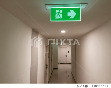Illuminated green emergency exit sign with running man symbol and arrow pointing right, mounted on ceiling in modern building corridor, safety guidance for evacuation and emergency preparedness 130435454