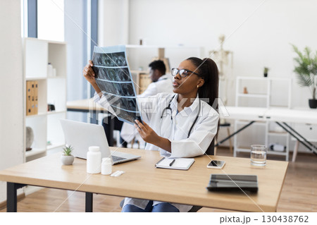 Confident young adult female doctor evaluating X-ray scans in office workspace. Medical professional focusing on diagnostics and patient records. Modern healthcare settings emphasizing professionalism 130438762