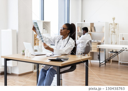 Young African woman doctor analyzing x-ray image at desk in contemporary clinic Young African woman doctor analyzing x-ray image at desk in contemporary clinic 130438763