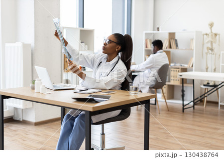 Young African woman doctor analyzing x-ray image at desk in contemporary clinic Young African woman doctor analyzing x-ray image at desk in contemporary clinic 130438764