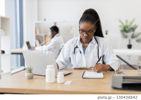 Young adult female doctor writing prescription using laptop and paper in office environment. Scene includes medication bottles and medical tools. Professional atmosphere demonstrating healthcare 130438842
