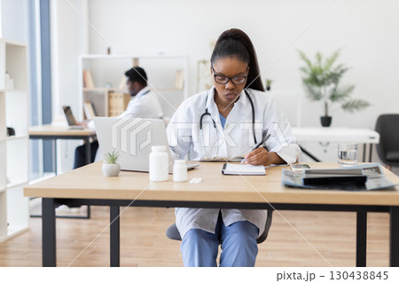 Young African American female healthcare worker in professional medical office writing prescription or report. Focused on task. Professional surroundings create an atmosphere of productivity and care. 130438845