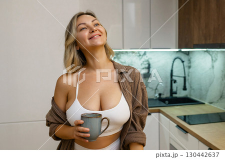 Modern kitchen: Young woman in a white crop top and open shirt stands with a mug in hand, enjoying a peaceful morning at home, relaxed and smiling. 130442637