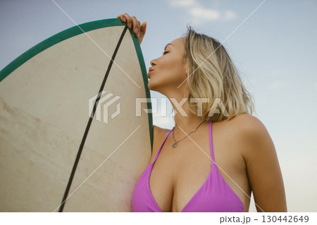 Blonde woman in purple bikini playfully kisses surfboard on sandy beach, summer sunset atmosphere, joyful and carefree vacation mood. Blonde woman in purple bikini playfully kisses surfboard on sandy beach, summer sunset atmosphere, joyful and carefree vacation mood. 130442649
