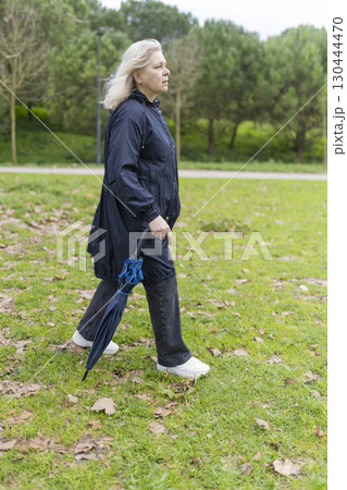 Senior woman walking in park with closed umbrella on cloudy day 130444470