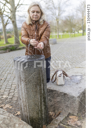 Senior woman washing hands in public park fountain on cold day Senior woman washing hands in public park fountain on cold day 130444473