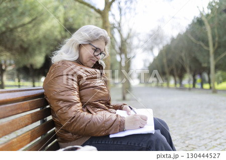 Senior woman writing on notepad while sitting on bench in park Senior woman writing on notepad while sitting on bench in park 130444527
