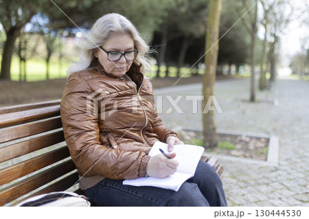 Senior woman writing in notebook while sitting on park bench 130444530