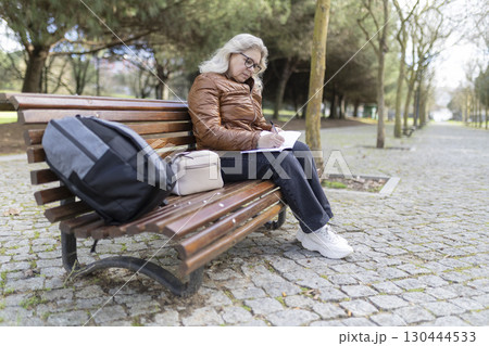 Senior woman writing notes in notebook while sitting on bench in park 130444533