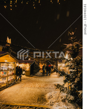 People strolling between wooden stalls adorned with twinkling Christmas lights, surrounded by softly falling snowflakes during magical winter evening in charming European city. Wooden market stalls 130448181