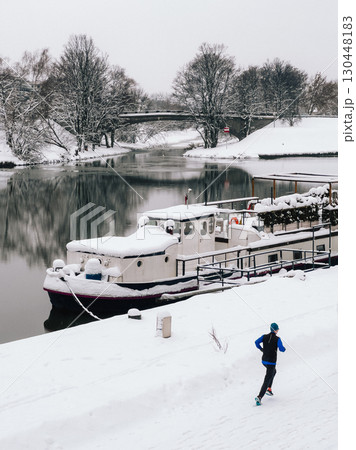 Man is jogging along snow covered riverbank, passing moored boat blanketed in snow, with bridge and snow laden trees in background during serene winter day. Male runs traversing snow covered riverbank 130448183