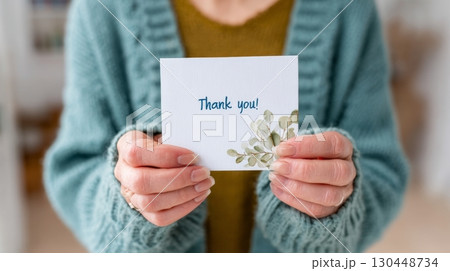 A person in a warm sweater holds a thank you card featuring a botanical design, expressing appreciation 130448734