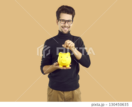 Young attractive man in transparent glasses, throws coins into yellow piggy bank in the form of pig. 130450635