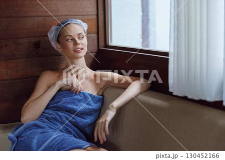 Relaxed woman wrapped in a blue towel with a hair towel turban, sitting by a window in a wooden spa room, enjoying a peaceful moment of self-care and calmness. Relaxed woman wrapped in a blue towel with a hair towel turban, sitting by a window in a wooden spa room, enjoying a peaceful moment of self-care and calmness. 130452166