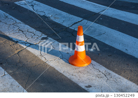 A traffic cone stands on a cracked crosswalk, casting shadows on the pavement in the morning light, indicating road work or caution 130452249