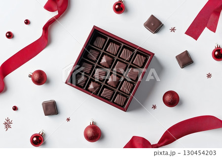 Christmas chocolate box mockup lying on a white surface surrounded by red ribbon, baubles and snowflakes 130454203