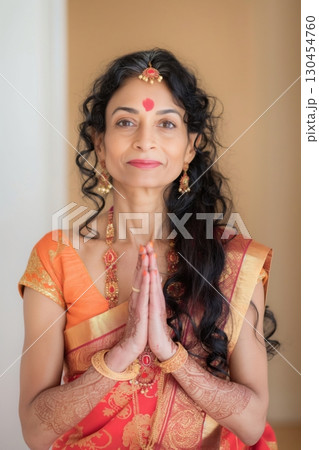 Elegant mature Indian woman with long curly black hair and a red dot on her forehead, wearing a red and orange sari and golden jewelry, holding her hands together in prayer Elegant mature Indian woman with long curly black hair and a red dot on her forehead, wearing a red and orange sari and golden jewelry, holding her hands together in prayer 130454760