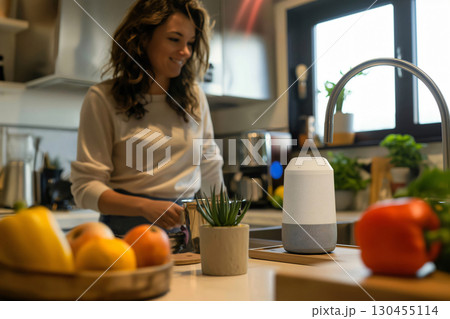 Woman happily uses a smart speaker while cooking in a modern kitchen with natural light, fresh fruits, and vegetables. Her smile shows enjoyment of the connected technology 130455114