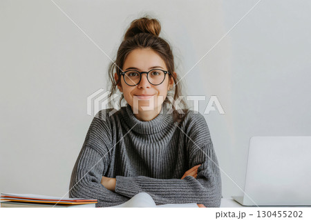 Young woman is sitting at a desk with her arms crossed, smiling, with a laptop and folders in front of her 130455202
