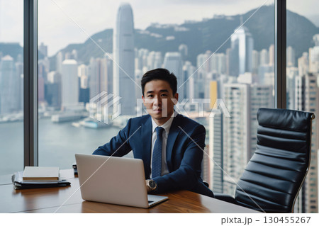 Young businessman is sitting at his desk in a modern office, working on his laptop with the cityscape of hong kong in the background 130455267