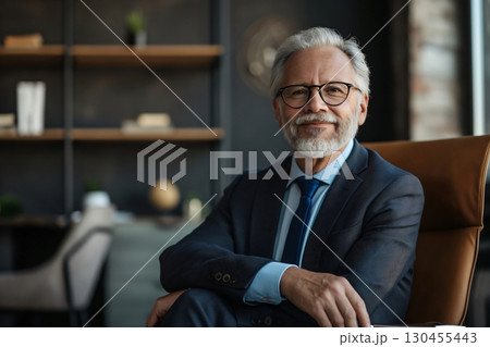 Mature businessman with a salt and pepper beard is sitting in his office chair, dressed in a suit and tie, looking confident 130455443