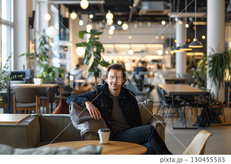 Young man is relaxing on a comfortable couch, enjoying a coffee break in a bustling modern cafe 130455583