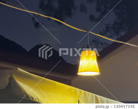 Yellow hanging lamp glowing in the evening with mountain silhouette in background. Illumination, electricity, night atmosphere, outdoor design and warm environment. 130457338