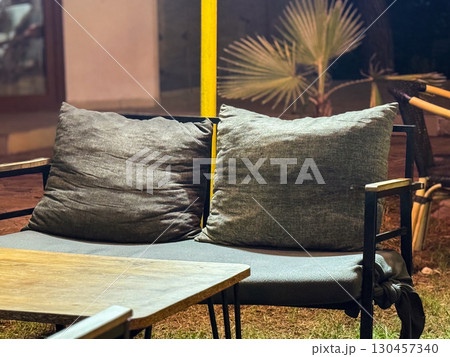 Outdoor sofa with large gray cushions placed near a table at night. Furniture, comfort, hospitality, leisure environment and relaxation space in a terrace. Outdoor sofa with large gray cushions placed near a table at night. Furniture, comfort, hospitality, leisure environment and relaxation space in a terrace. 130457340