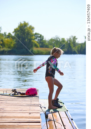 Young girl preparing for wakeboarding on wooden dock in summer lake Young girl preparing for wakeboarding on wooden dock in summer lake 130457409