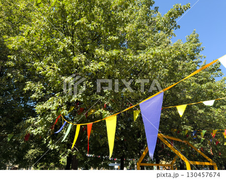 Colorful triangular flags hang across the trees in a vibrant outdoor setting. The clear blue sky and lush greenery add to the festive and joyful atmosphere, perfect for summer celebrations 130457674