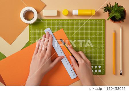 Hands cutting orange paper with a ruler and knife on a green mat. The process of creating art from paper. 130458023