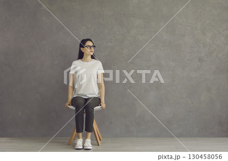 A young beautiful girl sits on a white stool in full growth, in a T-shirt, sports shoes. A young beautiful girl sits on a white stool in full growth, in a T-shirt, sports shoes. 130458056