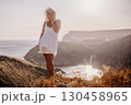 A blonde woman stands on a hill overlooking the ocean. She is wearing a white dress and she is enjoying the view. 130458965