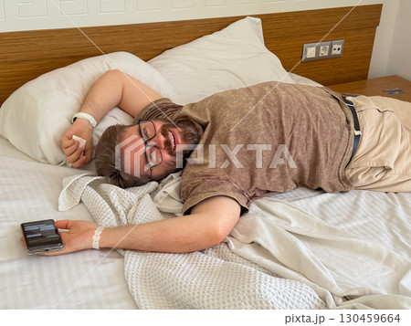 Smiling man lying on hotel bed with relaxed pose and casual mood. Vacation, leisure, connection and happiness of seasonal travel experience and everyday comfort. Smiling man lying on hotel bed with relaxed pose and casual mood. Vacation, leisure, connection and happiness of seasonal travel experience and everyday comfort. 130459664