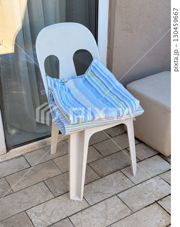 Plastic chair on a balcony with striped towel folded in morning light. Leisure, vacation, simplicity and everyday comfort during seasonal resort travel and accommodation. 130459667