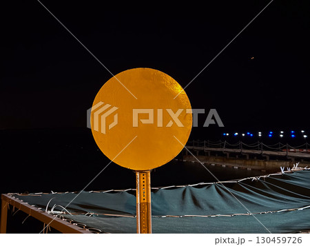 Round yellow sign illuminated against a dark night background near a pier. Minimal design, abstract geometry, symbol, outdoor installation and evening environment. 130459726
