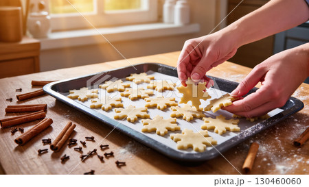 Hands placing raw snowflake-shaped cookies on baking tray in cozy kitchen. Christmas cookie preparation with cinnamon and cloves in warm morning light. Hands placing raw snowflake-shaped cookies on baking tray in cozy kitchen. Christmas cookie preparation with cinnamon and cloves in warm morning light. 130460060