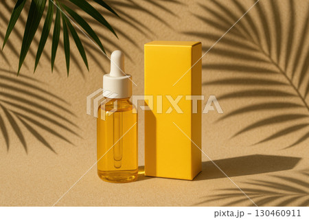 Yellow glass dropper bottle with matching box on sandy surface, palm leaves casting tropical shadows, modern skincare product display 130460911