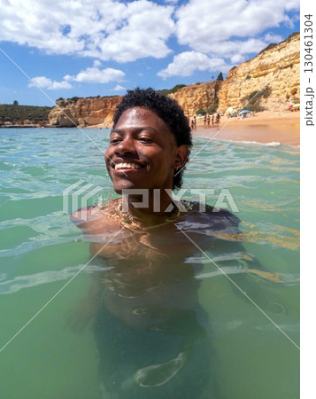 Young African American man swims in ocean in Algarve Portugal cliffs blue sky beach 130461304