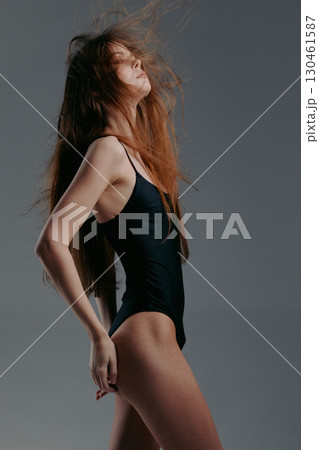 Young woman in black swimsuit posing with flowing hair on gray background, showcasing elegance and confidence in studio setting. 130461587
