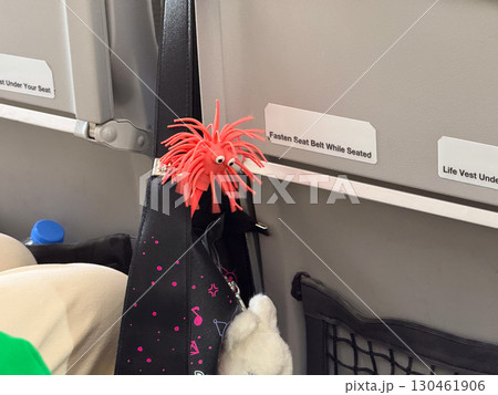 Red toy figure attached to passenger bag in airplane cabin interior. Travel accessory, playful decoration and lighthearted element of airline journey. 130461906