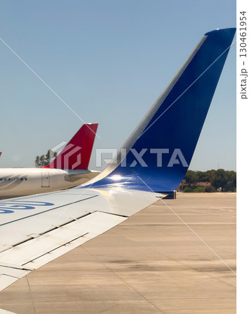 A close view of an airplane wing with blue tail fin and another red tail in the background. Aviation, technology, and the grounded preparation of aircraft before passenger flight. A close view of an airplane wing with blue tail fin and another red tail in the background. Aviation, technology, and the grounded preparation of aircraft before passenger flight. 130461954