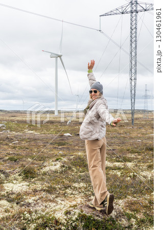 Woman in beige pants and a gray jacket raises her arms joyfully near wind turbines and power lines in Teriberka, Russia 130461961